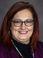 A woman with shoulder-length brown hair, glasses, and hoop earrings. She is smiling and wearing a dark blouse with a purple jacket.