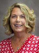 A woman with shoulder-length blonde curly hair, wearing a red and white patterned top, smiles in front of a gray background.