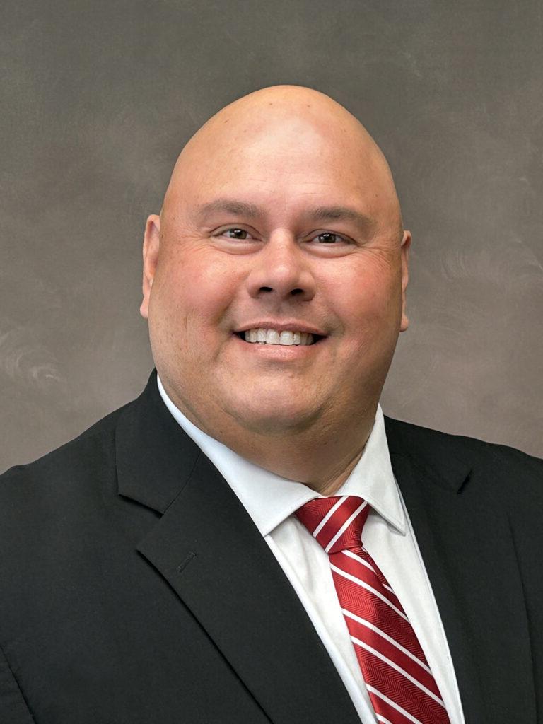 A bald man in a black suit jacket, white shirt, and red striped tie smiles at the camera against a neutral background.